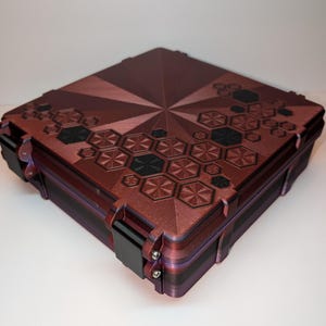 May include: A burgundy and black square box with a geometric design on the lid. The lid features a starburst pattern and black hexagonal shapes. The box has black latches and hinges, and a textured surface.