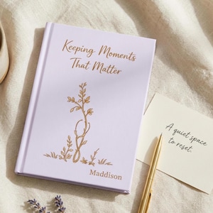 May include: A light purple hardcover journal with gold foil text reading Keeping Moments That Matter and the name Maddison. A gold line art illustration of a plant is centered. A gold pen and a card with the text A quiet space to reset.