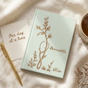 May include: A light green hardcover journal featuring a gold-colored botanical illustration and the name Bernadette printed on the cover. Next to the journal is a gold pen and a small white card with the text One day at a time written in brown.