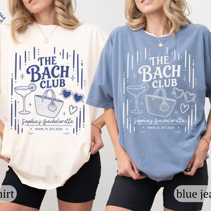 May include: Two t-shirts, one cream and one blue, with a graphic design. The design includes the text "THE BACH CLUB", a cocktail, a tote bag, heart-shaped sunglasses, and the text "Sophia's Bachelorette, Miami, FL EST 2026".