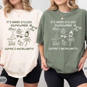 May include: Two t-shirts, one ivory and one moss green, with the text "IT'S BRIDE O'CLOCK SOMEWHERE August 2026" and "SOPHIA'S BACHELORETTE". The design includes illustrations of a cocktail, disco ball, sunglasses, champagne bottle, beach umbrella, rings, and a palm tree.