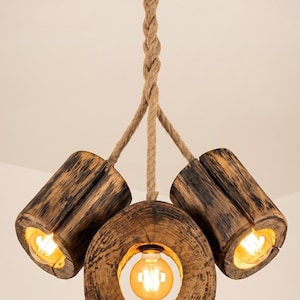 Puede incluir: Una lámpara de techo con tres segmentos de troncos de madera cilíndricos suspendidos por gruesas cuerdas de yute trenzadas desde un dosel circular de metal negro. Cada segmento de madera contiene una bombilla cálida tipo Edison.