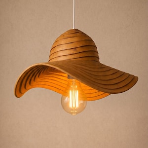 May include: A pendant light fitting featuring a wide, wavy, brown wooden shade shaped like a sun hat. The shade is constructed from stacked, curved layers of wood. A single clear glass light bulb with visible filaments hangs from a white cord.