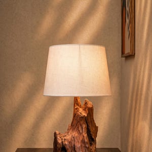 Könnte beinhalten: Eine Tischlampe mit einem Fuß aus natürlichem, strukturiertem braunem Treibholz. Die Lampe ist mit einem einfachen, cremeweißen, konischen Stofflampenschirm versehen, der ein weiches, warmes Licht abgibt und die komplexe Holzmaserung betont.