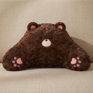 May include: A dark brown plush backrest pillow shaped like a bear. The pillow features a textured faux fur surface, two small rounded ears with pink inner accents, a cream-colored muzzle, and two extended arms with pink paw pad details.