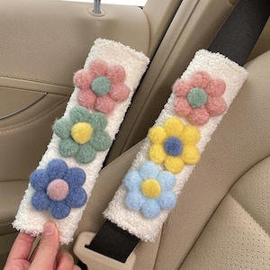 May include: Two white sherpa fabric seat belt covers featuring three-dimensional plush flowers. Each cover displays a unique arrangement of pastel-colored blossoms in shades of pink, green, yellow, and blue with contrasting centers.