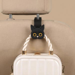 May include: A black car headrest hook featuring a stylized cat face with yellow eyes and a small pink nose. The hook is attached to a car seat headrest and is currently supporting the white, twisted rope handle of a small, rectangular white bag.