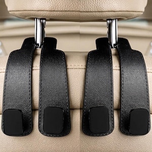 May include: A set of four black car seat headrest hooks made of textured material with visible stitching along the edges. Each hook features a flat, rectangular base and a curved top section designed to attach securely to the metal headrest posts.