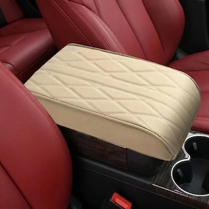 May include: A beige car centre console armrest cover featuring a diamond-stitched quilted pattern. The cover is positioned between red leather car seats and sits above a dark wood-grain trim panel with integrated circular cup holders.