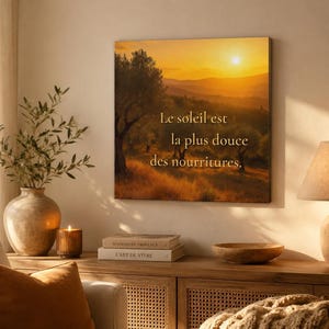 May include: A canvas print displays a sunset landscape with a tree and the text "Le soleil est la plus douce des nourritures." A vase with greenery, a lit candle, books, a wooden bowl, and a lamp are on a wooden cabinet.