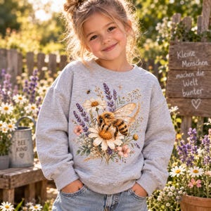 Könnte beinhalten: Ein hellgraues Sweatshirt mit Rundhalsausschnitt und einem zentralen Grafikdruck einer Honigbiene auf einer großen weißen Margerite, umgeben von violetten Lavendelzweigen, kleinen rosa Blüten und grünem Blattwerk, mit langen Ärmeln.