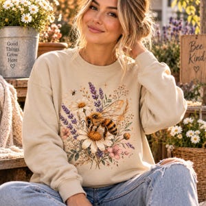 Könnte beinhalten: Ein beigefarbenes Sweatshirt mit Rundhalsausschnitt und einem zentralen Grafikdruck einer Honigbiene auf einem großen weißen Gänseblümchen, umgeben von violetten Lavendelzweigen, rosa Blüten und Weizenähren, verziert mit kleinen Sternen und einer Biene.