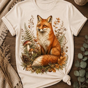 Könnte beinhalten: Cremefarbenes T-Shirt mit einer detaillierten Illustration eines Rotfuchses inmitten von Herbstlaub, einschließlich Pilzen, Tannenzapfen und Blättern. Der Fuchs hat einen flauschigen Schwanz und ist von Farnen und anderen Pflanzen umgeben. Das T-Shirt ist unten geknotet.