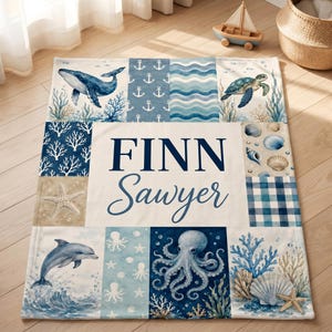 May include: A rectangular blanket featuring a nautical theme with a central white panel displaying the name FINN Sawyer in dark blue text. The surrounding panels showcase illustrations of a whale, sea turtle, dolphin, octopus, shells, and anchors.