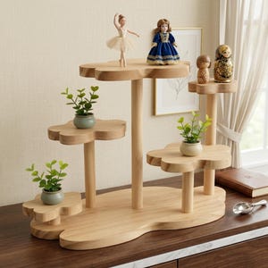 May include: A multi-tiered wooden display stand with flower-shaped platforms at varying heights. The stand holds three small potted green plants, a ballerina figurine, a doll in a blue dress, a small wooden figure, and a decorative nesting doll.