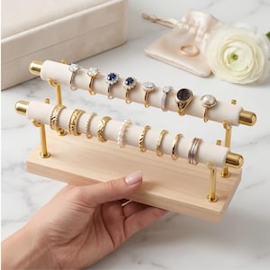 May include: A two-tier ring display stand featuring gold metal rods with soft beige velvet padding, mounted on a light wood base. The stand holds sixteen various rings, including diamond, sapphire, pearl, and gold bands, arranged in two rows.