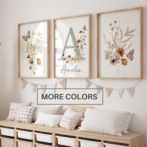May include: Three framed wall art prints featuring botanical illustrations of wildflowers and butterflies. The center print displays a large letter A with the name Amelia below. A wooden storage bench with white bins sits below the art.