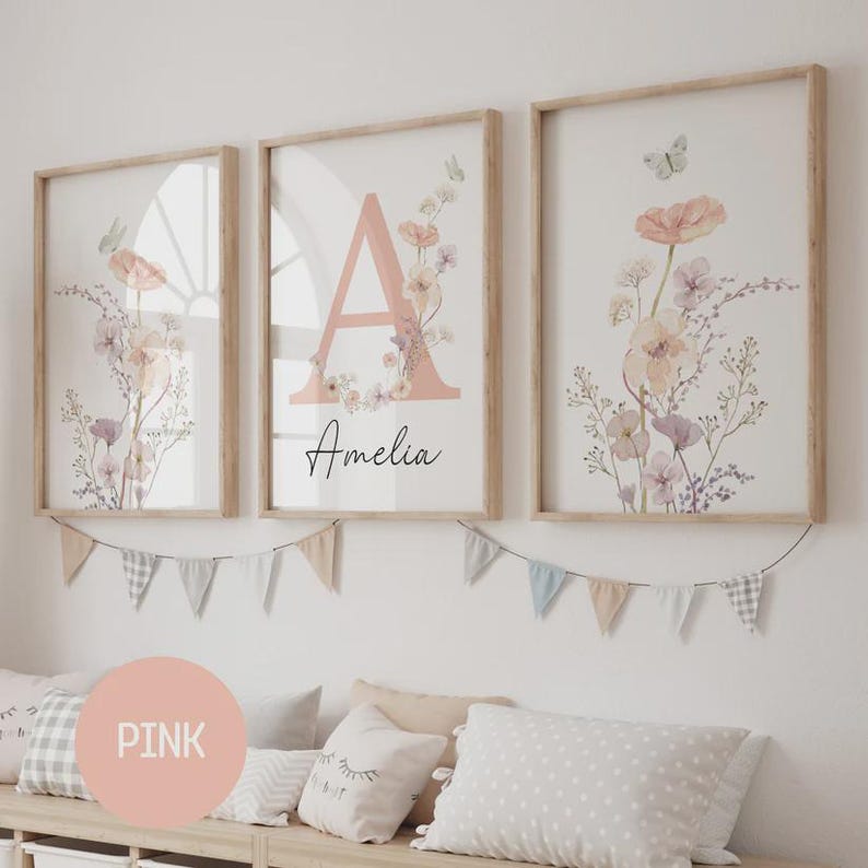 May include: Three framed floral art prints. The prints feature watercolor flowers in soft pinks, purples, and creams. One print has a large pink letter "A" with the name "Amelia" below. The frames are light wood.