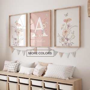 May include: Three framed wall art prints featuring watercolor floral illustrations and butterflies. The center print displays a large white letter A and the name Amelia. A string of fabric bunting hangs below the frames.