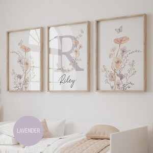 May include: Three framed floral art prints. The prints feature watercolor flowers in peach, lavender, and cream tones, with butterflies. The center print has a large lavender letter "R" with the name "Riley" below.