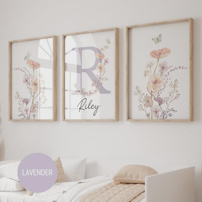 May include: Three framed floral art prints. The prints feature watercolor flowers in peach, lavender, and cream tones, with butterflies. The center print has a large lavender letter "R" with the name "Riley" below.