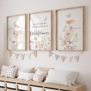 May include: Three framed wall art prints featuring watercolor illustrations of wildflowers, butterflies, and the text IN A FIELD OF ROSES SHE IS A Wildflower. The prints are displayed in light wood frames above a storage unit with decorative bunting.