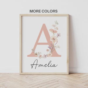 May include: Framed print featuring a large, blush-colored letter "A" adorned with watercolor flowers and a butterfly. The name "Amelia" is written in cursive below the letter. The words "MORE COLORS" are at the top.
