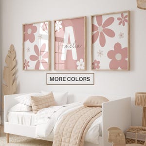 May include: Three rectangular wall art prints in light wooden frames featuring a dusty pink and white floral pattern. The center print displays a large white letter A with the name Amelia written in a thin script font. A box says MORE COLORS.