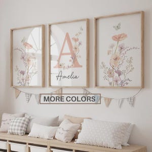 May include: Three framed floral art prints with a light wood frame. The prints feature a large letter "A" with the name "Amelia" and floral and butterfly accents. A banner below reads "MORE COLORS".