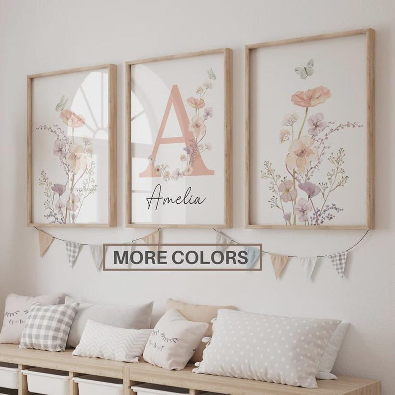 May include: Three framed floral art prints with a light wood frame. The prints feature a large letter "A" with the name "Amelia" and floral and butterfly accents. A banner below reads "MORE COLORS".