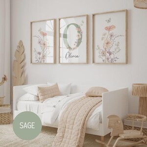 May include: Three framed floral prints with a light wood finish. The prints feature watercolor flowers in shades of pink, peach, and purple, with butterflies. The center print has a large letter "O" with the name "Olivia" below. The prints are above a white bed.