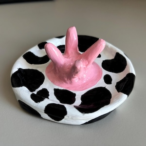 Può includere: Un piattino rotondo in ceramica con base bianca e macchie nere che ricordano il manto di una mucca. Al centro del piatto, una scultura rosa a tre punte simile a una mammella di mucca si estende verso l'alto da una base circolare rosa.