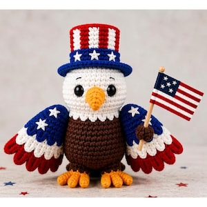 May include: Crocheted bald eagle figurine wearing a red, white, and blue Uncle Sam hat and holding a miniature American flag. The eagle has brown and white body, blue and white wings, and orange beak and feet. A patriotic themed decoration.