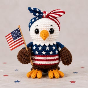 Op de afbeelding: Een gehaakte Amerikaanse zeearend pop met een blauw-wit sterrenpatroon shirt en een rood, wit en blauw hoofdband. De adelaar houdt een kleine Amerikaanse vlag vast. De pop heeft bruine vleugels en gele voeten.