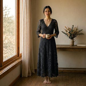 Può includere: Un abito in pizzo grigio scuro con scollo a V e maniche a tre quarti. L'abito lungo presenta un motivo floreale. Una persona tiene in mano una tazza beige. Lo sfondo include una finestra, tende e un tavolo di legno con un vaso di fiori.