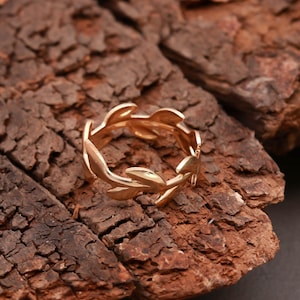 Op de afbeelding: Een goudkleurige ring met een doorlopend ontwerp van gestileerde bladeren. De ring is geplaatst tegen een getextureerde, donkerbruine achtergrond, waardoor een contrast ontstaat dat de ingewikkelde details van de ring benadrukt. Het ontwerp van de ring suggereert een op de natuur geïnspireerd thema.