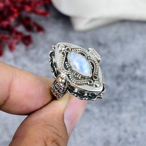 Op de afbeelding: Versierde zilveren ring met een ovale, iriserende maansteen. De ring heeft gedetailleerde filigraan en krullen rond de steen en op de band. De zetting is een gedetailleerd, scharnierend medaillonontwerp. De ring wordt door een hand vastgehouden.