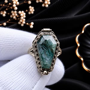 May include: A silver ring featuring a coffin-shaped setting with a faceted green moss agate stone. The metal frame has a textured, beaded border detail surrounding the gemstone. The ring band is split into two thin, polished silver segments.