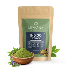 Peut inclure: Un paquet de poudre d'indigo Mohana Wellness, une teinture capillaire naturelle. Le paquet est beige avec une étiquette verte et le texte "INDIGO POWDER". Un bol en bois rempli de poudre verte et des brins de feuilles et de fleurs sont au premier plan. Contient 100g.