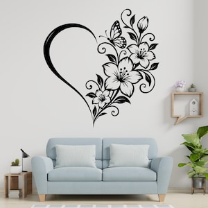 May include: A black wall decal featuring a heart outline on the left side, with a detailed floral arrangement of lilies, leaves, and a butterfly extending along the right side. The design is composed of clean, solid black lines and shapes.