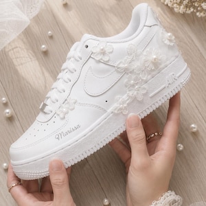 Könnte beinhalten: Ein weißer Sneaker mit Schnürverschluss, seitlichem Swoosh-Logo und dem Namen Marissa auf der Zehenkappe. Der Schuh ist mit weißen floralen Spitzenapplikationen und kleinen weißen Perlen an der Seite und im Fersenbereich verziert.