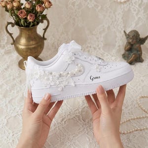 Może przedstawiać: Biały sneaker z koronkową aplikacją kwiatową z boku, ozdobiony małymi białymi perłami wzdłuż logo swoosh. But posiada białą wstążkę zamiast sznurowadła oraz imię Grace nadrukowane czarną czcionką na bocznej części podeszwy.