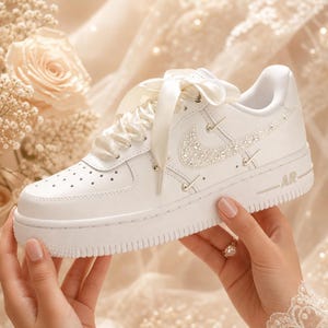 May include: White leather sneaker with a jeweled Nike swoosh and satin ribbon laces. The shoe features a classic design with perforations on the toe box and the word "AR" on the side. A perfect bridal shoe.