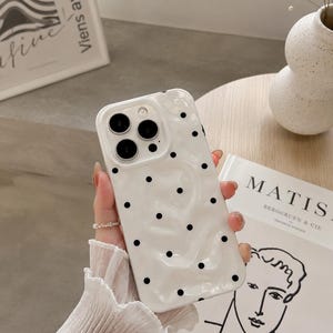 May include: White phone case with a glossy finish and a scattered black polka dot pattern. The case has a camera cutout with four lenses. The phone case is held in a hand.