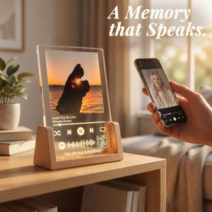 May include: A clear acrylic photo display with a wooden base. The display shows a sunset silhouette image and song details. A smartphone displays a photo of a woman. Text on the display reads "A Memory that Speaks."
