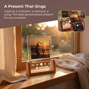 Puede incluir: Una pantalla fotográfica de acrílico transparente con una base de madera, que presenta una imagen de silueta de puesta de sol. La pantalla incluye controles de música y el texto "A Present That Sings". Un teléfono inteligente muestra la misma imagen y controles de música.