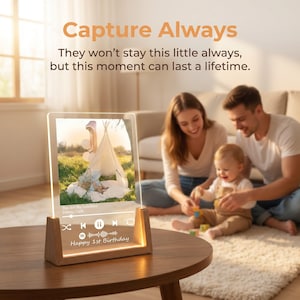 Può includere: Una targa rettangolare in acrilico trasparente con base in legno, raffigurante una fotografia di un bambino in una tenda, un'interfaccia di lettore musicale con il testo "You Are My Sunshine" e la scritta "Happy 1st Birthday".