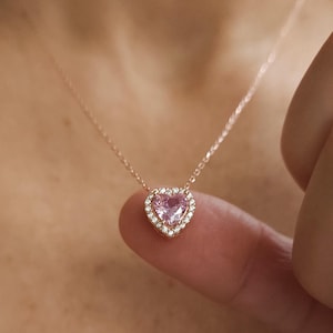 Könnte beinhalten: Eine zarte Halskette aus Roségold mit einem herzförmigen Anhänger, der einen rosafarbenen Edelstein mit kleineren, klaren Edelsteinen umrahmt.