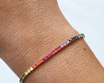 May include: A rainbow-colored tennis bracelet with a spectrum of gemstones. The bracelet features a row of small, round gemstones in a gradient of colors, from yellow to red to purple. The bracelet is worn on a wrist.