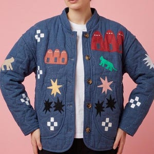 May include: A quilted denim jacket in a medium blue hue, adorned with colorful appliqués. The jacket features a round collar, button closure, and various shapes, including stars, houses, and a green animal. The jacket is worn over a white t-shirt.